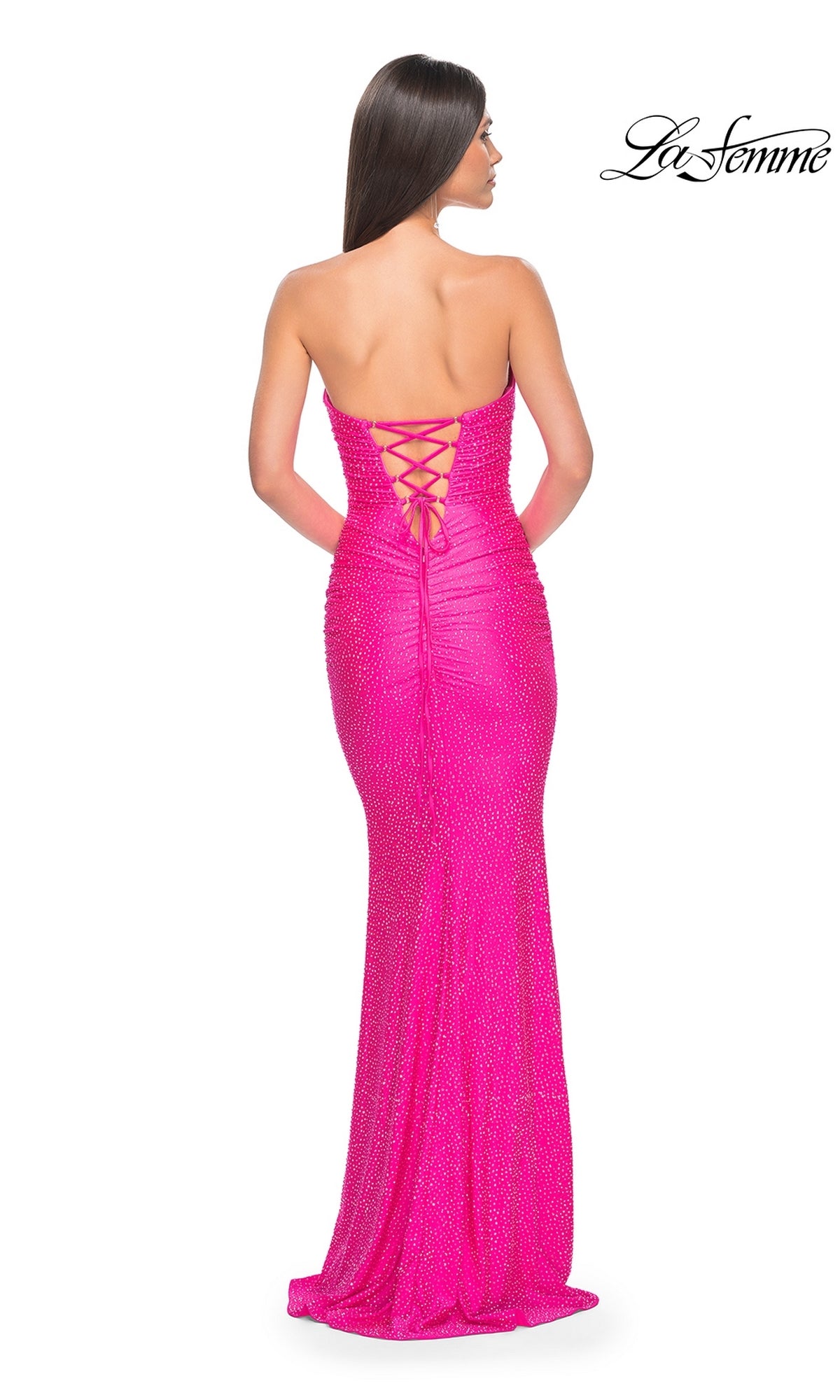 A woman poses with her back to the camera in a neon pink strapless long prom dress featuring a lace-up back and subtle beading.